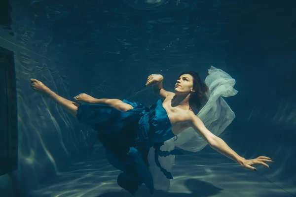 cute sporty female swims underwater as a free diver in a blue evening dress alone