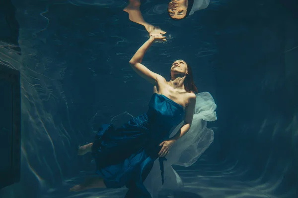 cute sporty female swims underwater as a free diver in a blue evening dress alone