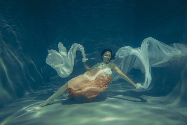 elegant slender girl swims underwater like a free diver in a white evening dress with beautiful fabric