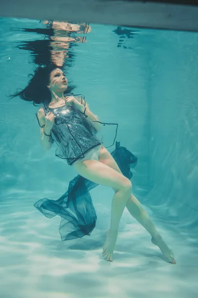 stock image beautiful model girl swims under water in a t-shirt in gray sequins and a transparent fashionable PVC raincoat