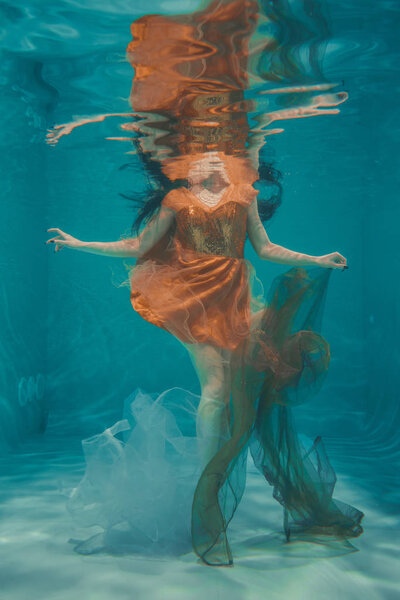 beautiful model girl swims underwater in orange dress and enjoys relaxation and lack of stress