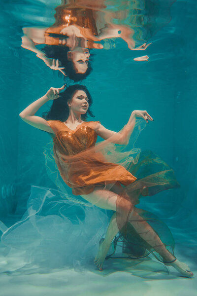 beautiful model girl swims underwater in orange dress and enjoys relaxation and lack of stress