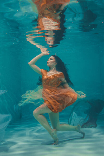 beautiful model girl swims underwater in orange dress and enjoys relaxation and lack of stress