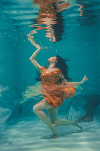 beautiful model girl swims underwater in orange dress and enjoys relaxation and lack of stress