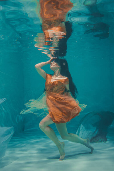 beautiful model girl swims underwater in orange dress and enjoys relaxation and lack of stress