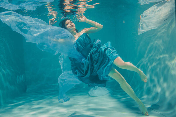 beautiful model girl with long black hair swims underwater in evening blue dress and enjoys relaxation and lack of stress