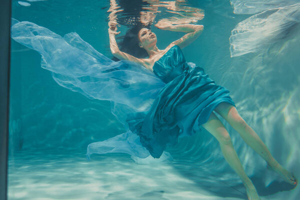 beautiful model girl with long black hair swims underwater in evening blue dress and enjoys relaxation and lack of stress
