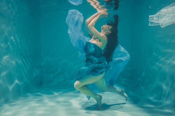 beautiful model girl with long black hair swims underwater in evening blue dress and enjoys relaxation and lack of stress