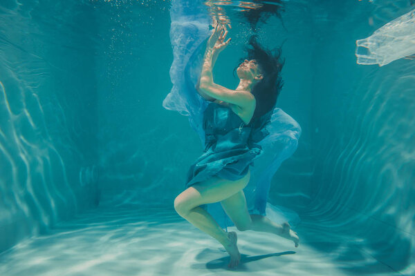 beautiful model girl with long black hair swims underwater in evening blue dress and enjoys relaxation and lack of stress
