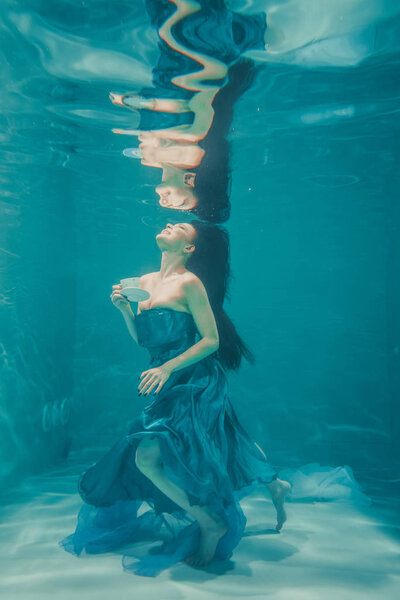 funny girl having fun and drinking soothing tea for Breakfast while meditatively swimming underwater in a dress
