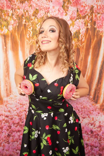 美丽的年轻女孩站在黑色别针礼服和有乐趣玩釉面粉红色甜甜圈 — 图库照片