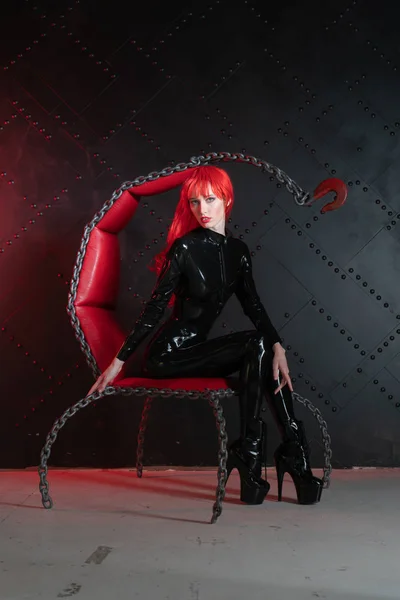 危险的残酷恋物癖女孩与红头发摆姿势黑色的 Bdsm 乳胶衣服在椅子上的链在虐待狂的风格 — 图库照片
