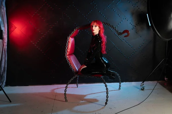 サドマゾヒズムのスタイルのチェーンから椅子に黒い Bdsm ラテックス服でポーズをとって赤い髪の危険な残酷なフェチ少女 — ストック写真