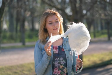 Beyaz kız büyüleyici bir kot ceket baharda Park içeri girer ve pembe pamuk şeker, sabah güneş ışığı gülümsüyor yiyor