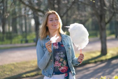 Beyaz kız büyüleyici bir kot ceket baharda Park içeri girer ve pembe pamuk şeker, sabah güneş ışığı gülümsüyor yiyor