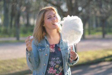 Beyaz kız büyüleyici bir kot ceket baharda Park içeri girer ve pembe pamuk şeker, sabah güneş ışığı gülümsüyor yiyor