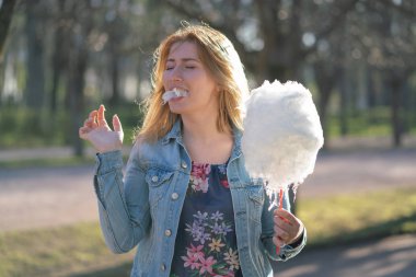 Beyaz kız büyüleyici bir kot ceket baharda Park içeri girer ve pembe pamuk şeker, sabah güneş ışığı gülümsüyor yiyor