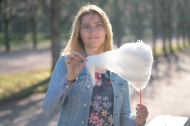 Beyaz kız büyüleyici bir kot ceket baharda Park içeri girer ve pembe pamuk şeker, sabah güneş ışığı gülümsüyor yiyor