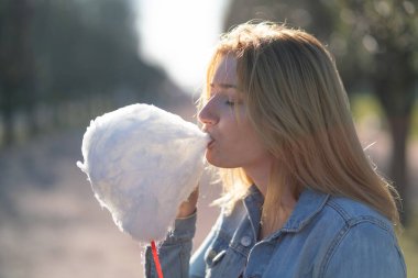 Beyaz kız büyüleyici bir kot ceket baharda Park içeri girer ve pembe pamuk şeker, sabah güneş ışığı gülümsüyor yiyor