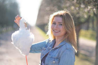 Beyaz kız büyüleyici bir kot ceket baharda Park içeri girer ve pembe pamuk şeker, sabah güneş ışığı gülümsüyor yiyor