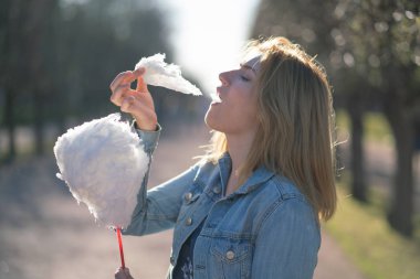Beyaz kız büyüleyici bir kot ceket baharda Park içeri girer ve pembe pamuk şeker, sabah güneş ışığı gülümsüyor yiyor