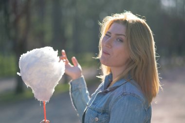 Beyaz kız büyüleyici bir kot ceket baharda Park içeri girer ve pembe pamuk şeker, sabah güneş ışığı gülümsüyor yiyor