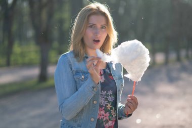 Beyaz kız büyüleyici bir kot ceket baharda Park içeri girer ve pembe pamuk şeker, sabah güneş ışığı gülümsüyor yiyor