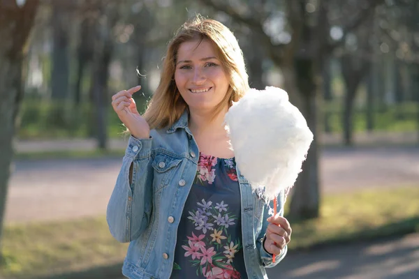 Beyaz kız büyüleyici bir kot ceket baharda Park içeri girer ve pembe pamuk şeker, sabah güneş ışığı gülümsüyor yiyor