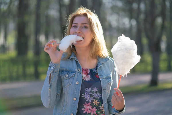 Beyaz kız büyüleyici bir kot ceket baharda Park içeri girer ve pembe pamuk şeker, sabah güneş ışığı gülümsüyor yiyor