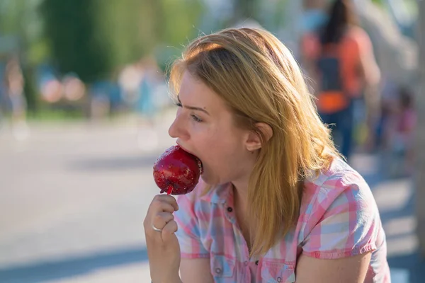 pretty young woman eating and biting red caramel apple in the park in the sunny day