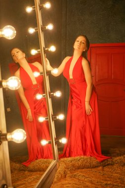 bir çerçeve ampuller ile büyük bir aynada yakınındaki bir kürk halı üzerinde kırmızı uzun akşam elbiseli çekici güzel kız