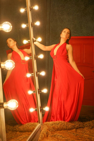 charming beautiful girl in a red long evening dress on a fur rug near a huge mirror in a frame with bulbs