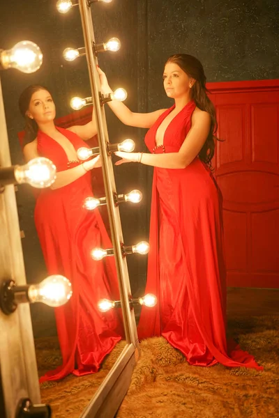 迷人美丽的女孩在一个红色的长晚礼服在毛皮地毯附近一个巨大的镜子在一个框架与灯泡 — 图库照片