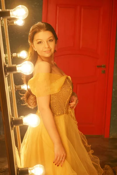 穿着金色晚礼服的美丽年轻女孩站在一个大镜子附近的皮草地毯上 在一个有灯光的框架里 看着她的倒影 — 图库照片