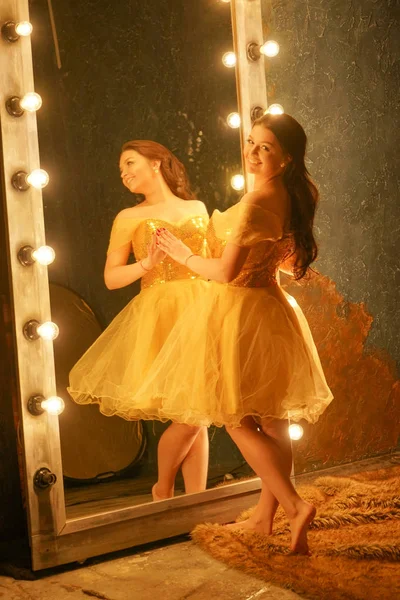 ゴールドのイブニング ドレスの美しい少女ライトとフレームに大きな鏡の近くの毛皮の敷物の上に立つし 彼女の反射に見える — ストック写真