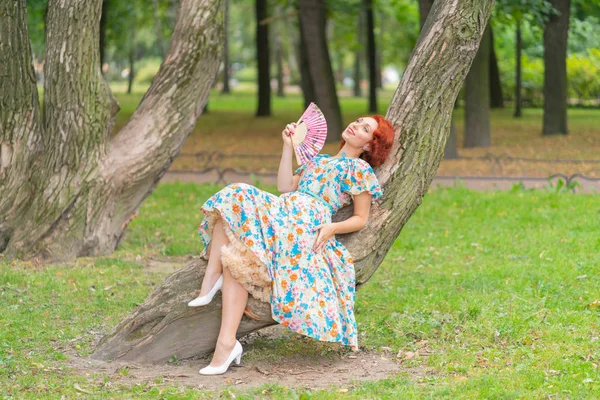 レトロなスタイルでヴィンテージのドレスで公園の木に彼女の手に扇を持ってポーズをとって赤髪の可愛い女の子 — ストック写真