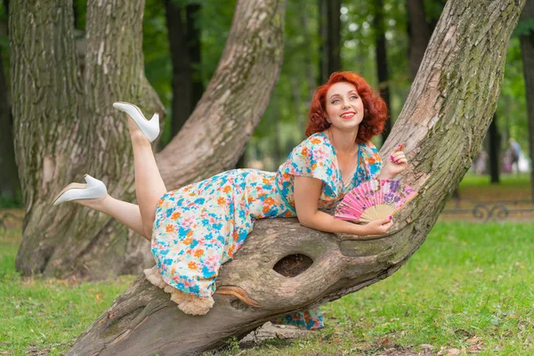 レトロなスタイルでヴィンテージのドレスで公園の木に彼女の手に扇を持ってポーズをとって赤髪の可愛い女の子 — ストック写真