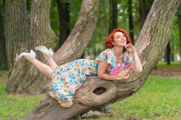 レトロなスタイルでヴィンテージのドレスで公園の木に彼女の手に扇を持ってポーズをとって赤髪の可愛い女の子 — ストック写真