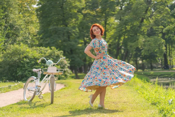 夏の日で都市公園でスポーツのファッショナブルな散歩で白のビンテージ自転車との生活を楽しんでいるレトロなドレスに赤い髪のかわいい白人の女の子 — ストック写真