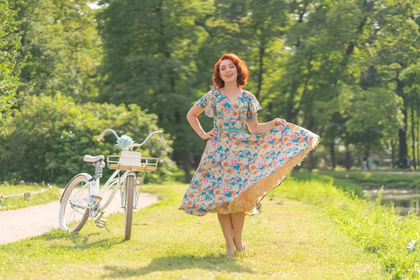 夏の日で都市公園でスポーツのファッショナブルな散歩で白のビンテージ自転車との生活を楽しんでいるレトロなドレスに赤い髪のかわいい白人の女の子 — ストック写真