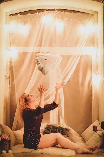 charming teenager girl in a red dress with sequins sits on the window sill with a white curtain and yellow round lamp light bulbs