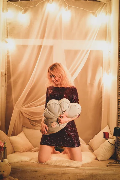 charming teenager girl in a red dress with sequins sits on the window sill with a white curtain and yellow round lamp light bulbs