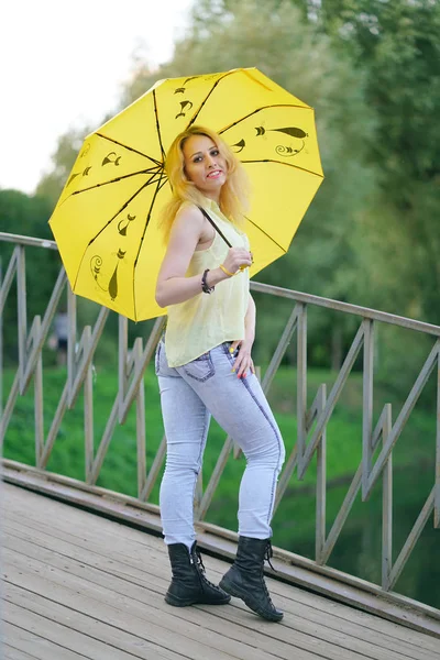 cute girl in yellow shirt and jeans walking on the bridge with a bright umbrella at the evening
