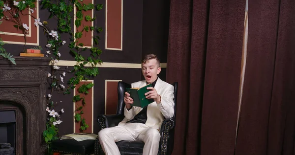 bored teen student guy in white business suit sitting in a chair with a book by the fireplace