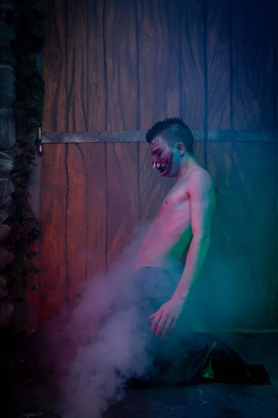 scary vampire male with huge teeth and bloody body as halloween body art standing in the night smoke alone