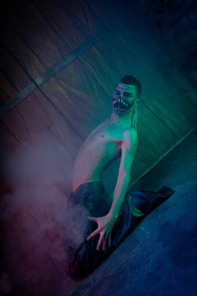 scary vampire male with huge teeth and bloody body as halloween body art standing in the night smoke alone