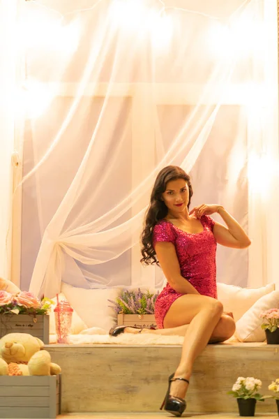 pretty girl in pink sequins dress having rest on evening window sill with light bulbs, pillows, flowers and toys