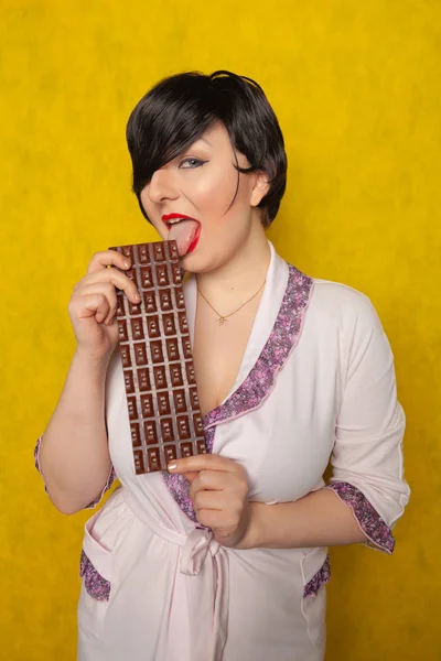 ピンクのバスローブと彼女の手で黄色の背景上にチョコレートの巨大な棒を持って立っているサイズの女性プラス — ストック写真