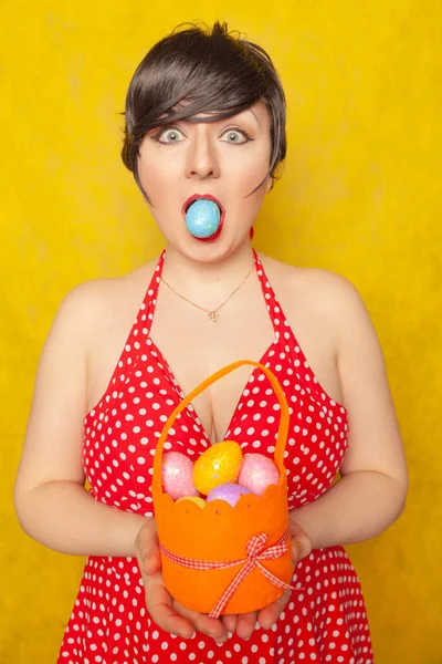 cute girl with short black hair dressed in a red retro polka dot dress posing with orange basket of colored eggs for Easter on a yellow solid background in the Studio