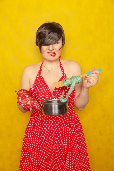 plump girl in a red polka-dot dress stands with an empty pan and a centimeter tape on a spoon, wants to eat, but diets for health and figure